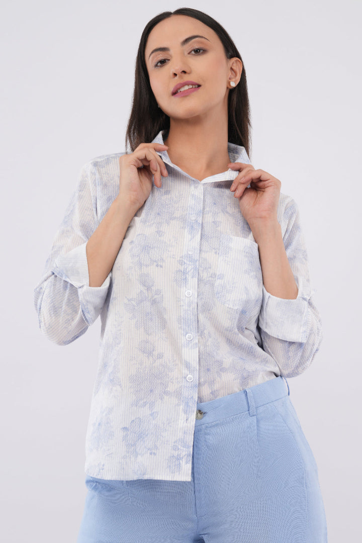 A woman wearing a light blue and white floral patterned button-up shirt with light blue trousers, posing against a solid light gray background.